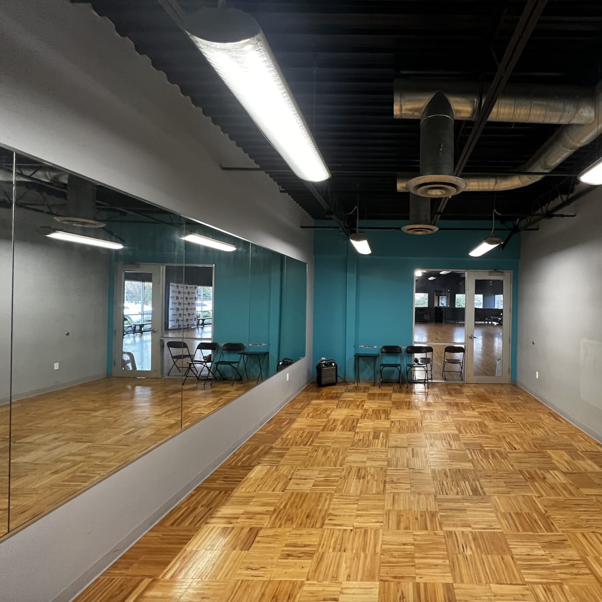 A spacious room with a wooden floor, two mirrors on one wall, and several chairs arranged near a blue accent wall.