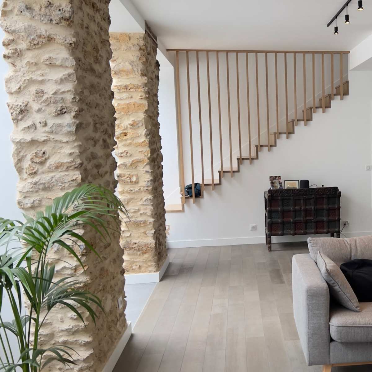 A modern living room featuring a gray sofa, a wooden staircase with a minimalist railing, and exposed stone walls beside a decorative cabinet.