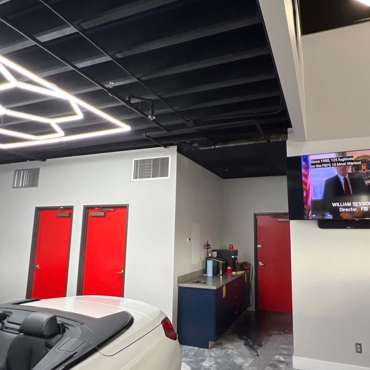 An interior space with a white convertible car in the foreground, red doors on the left, and a television mounted on the wall displaying a news segment.