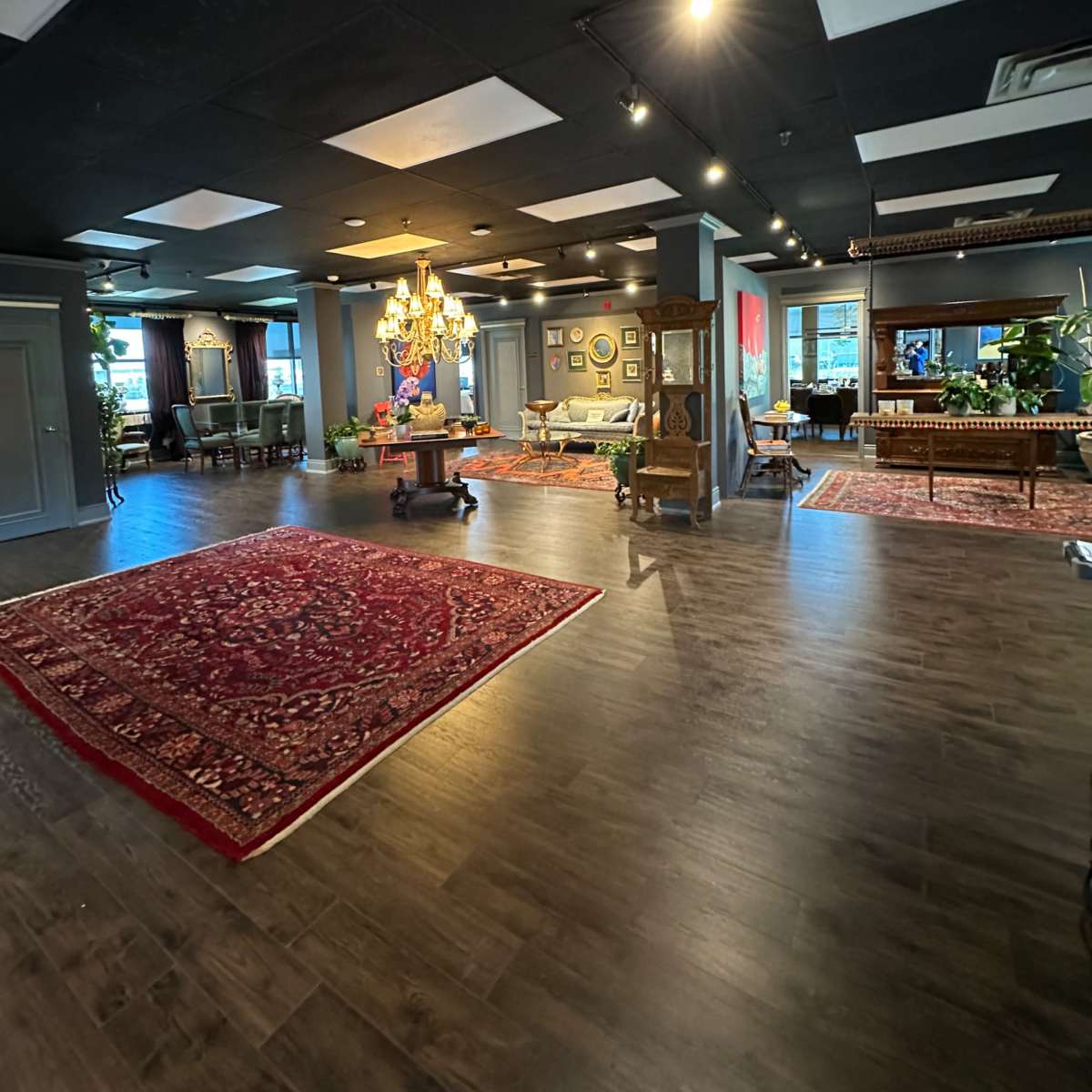 A spacious, elegantly designed room with dark wood flooring, decorative rugs, and various pieces of furniture including a chandelier and art on the walls.