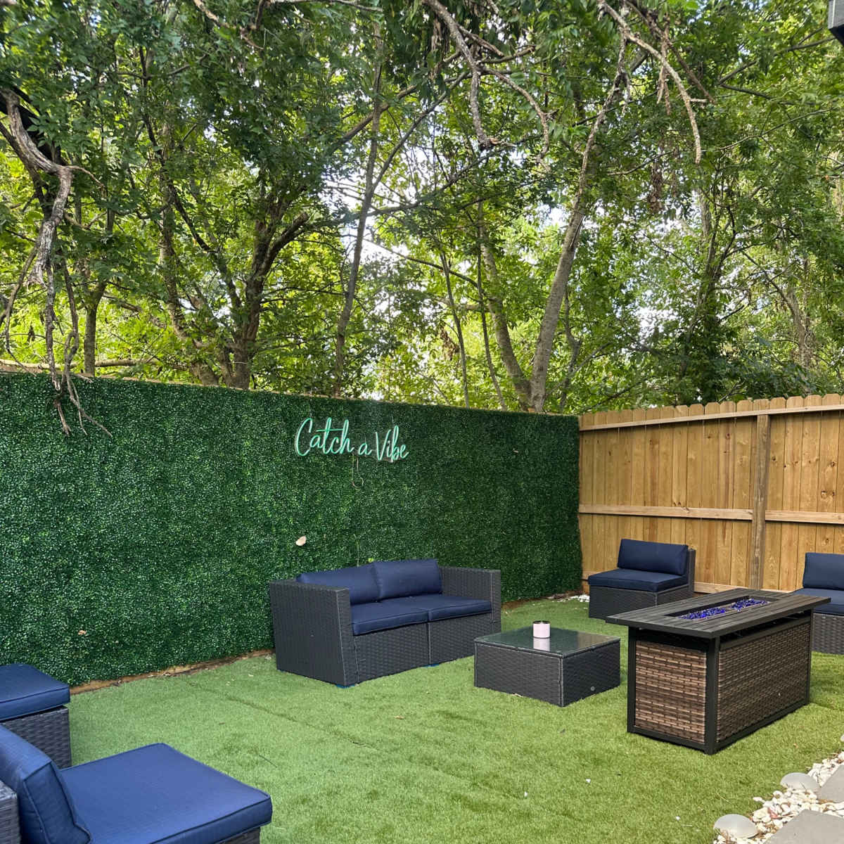 A outdoor seating area with dark wicker furniture arranged on a green artificial lawn, surrounded by trees and a wooden fence.