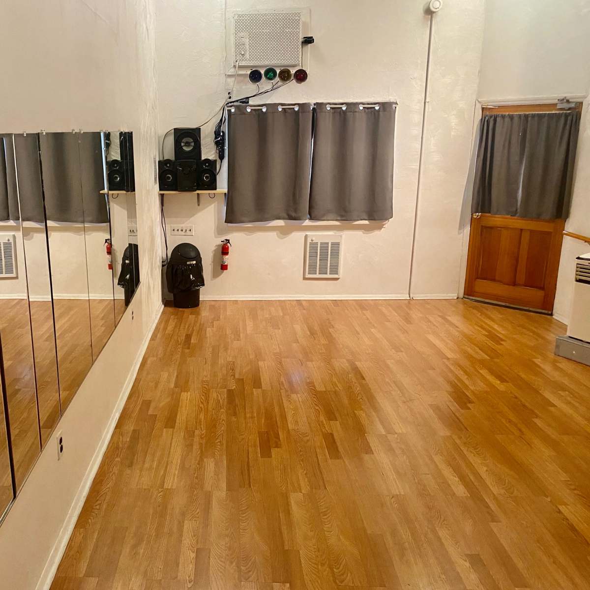 A dance studio with wooden flooring, mirrors on one wall, gray curtains, a door, and some audio equipment.