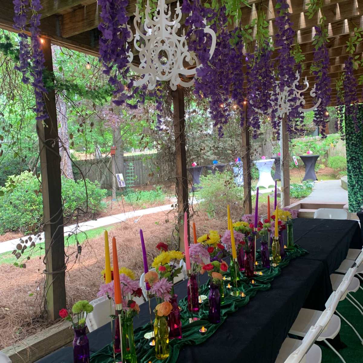 A long table adorned with colorful candles in glass vases is set under a pergola decorated with purple flowers and white chandeliers.