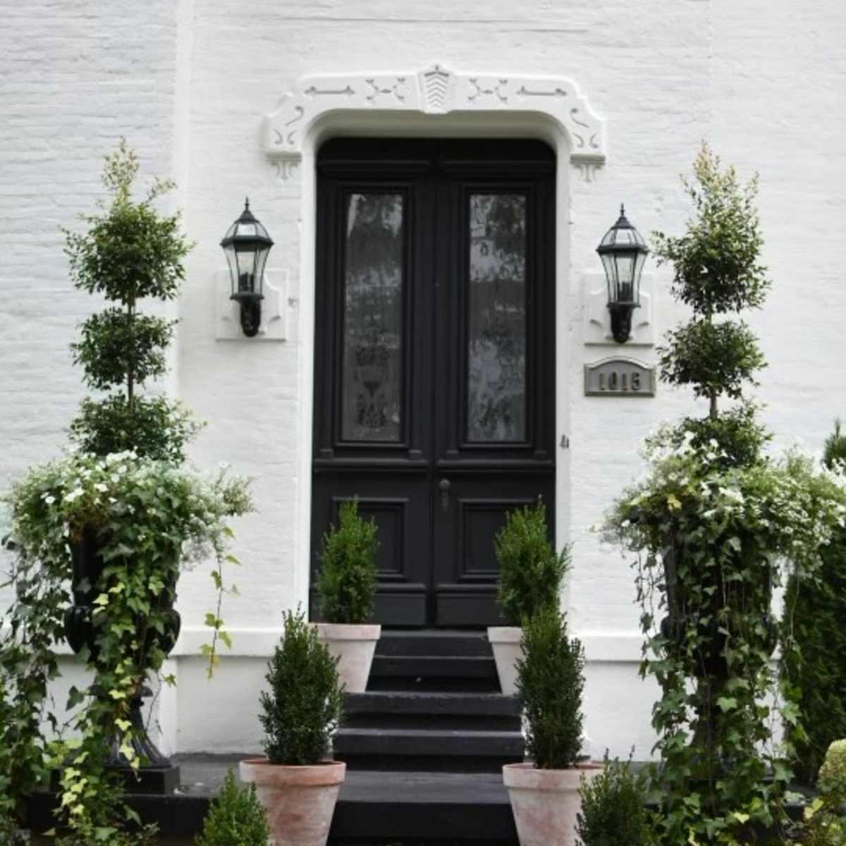 A white house features a dark front door flanked by potted plants on either side, with a stairway leading up to the entrance.