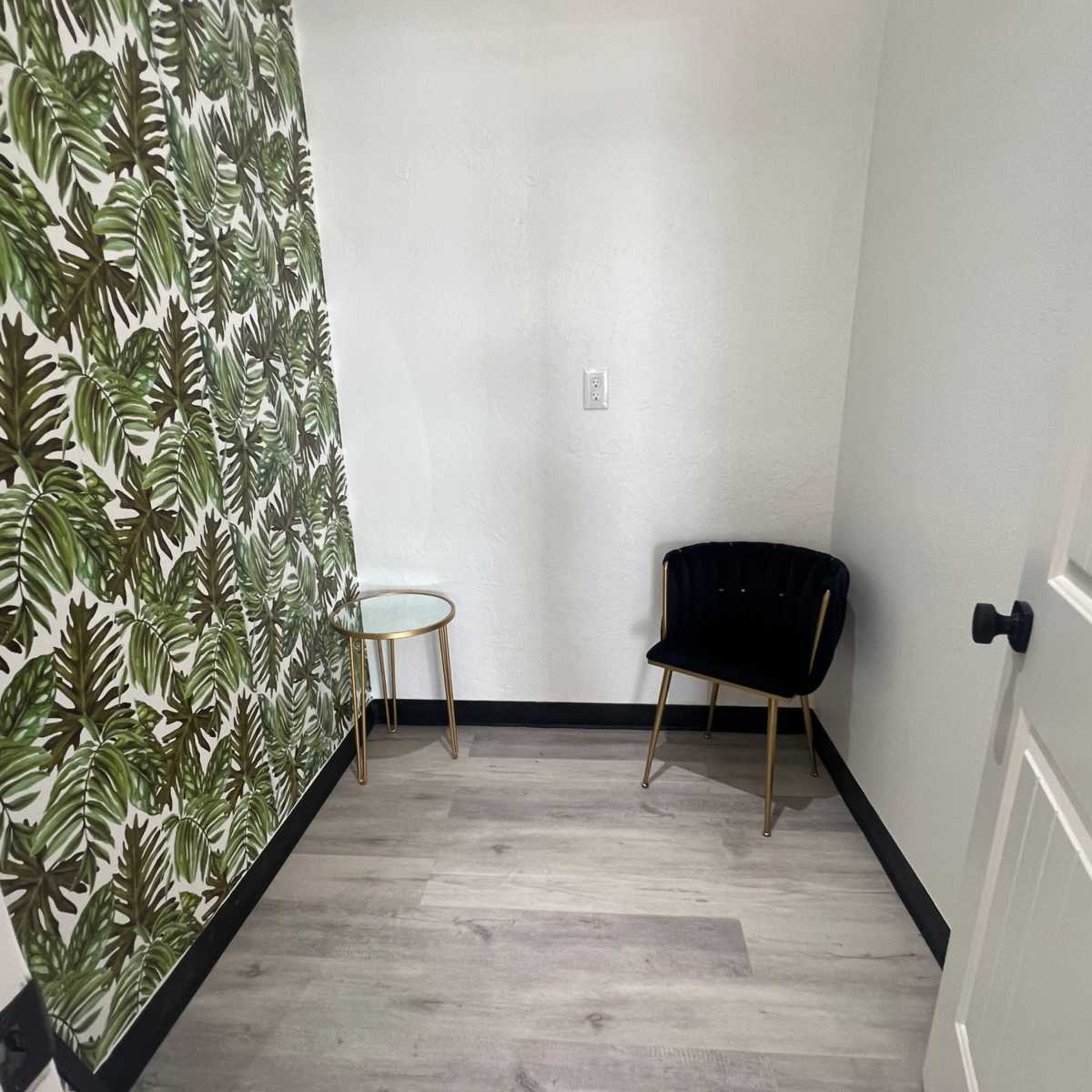 A small corner of a room featuring a patterned green leaf wallpaper, a black chair, and a small side table on a light wood floor.