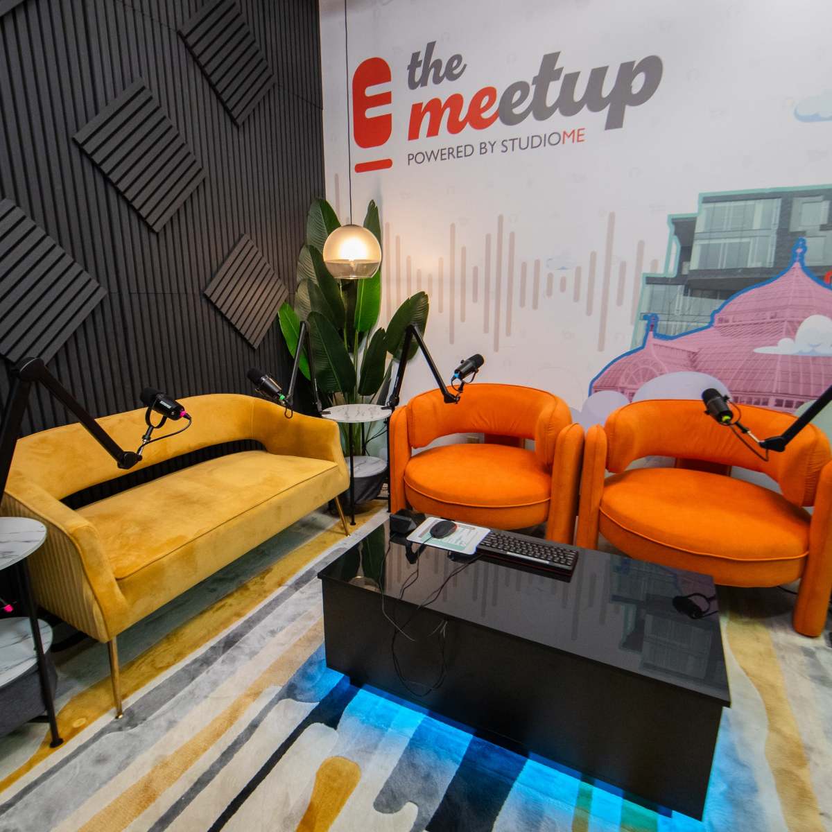 A modern podcast studio featuring two brightly colored chairs, a yellow couch, round side tables, and microphones set up on stands, against a backdrop with a mural of urban architecture.