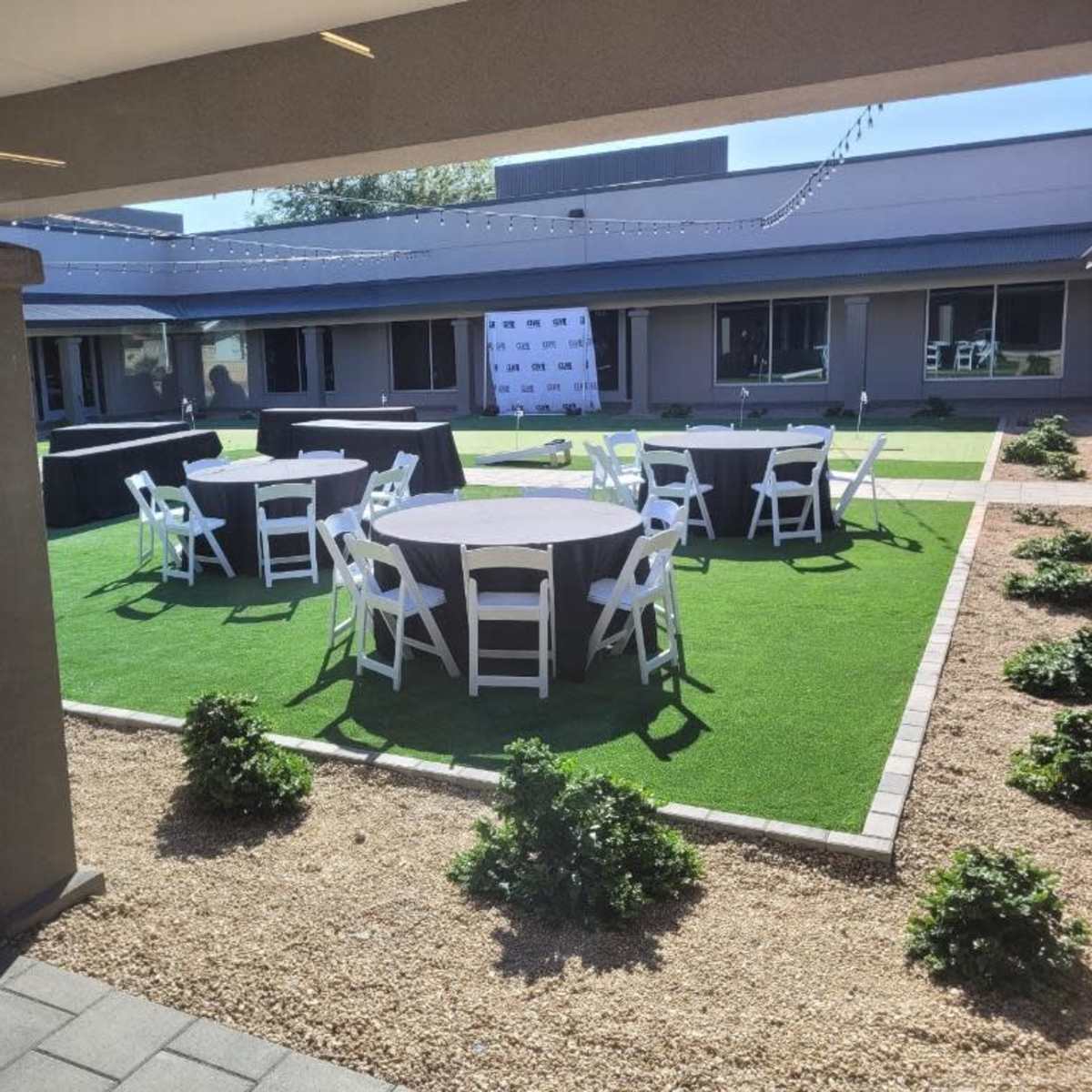 An outdoor event space with several round tables set up on grass and surrounded by hedges.