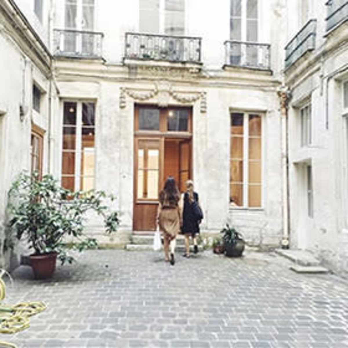 Two women walk toward a wooden door in a courtyard with stone paving and potted plants.