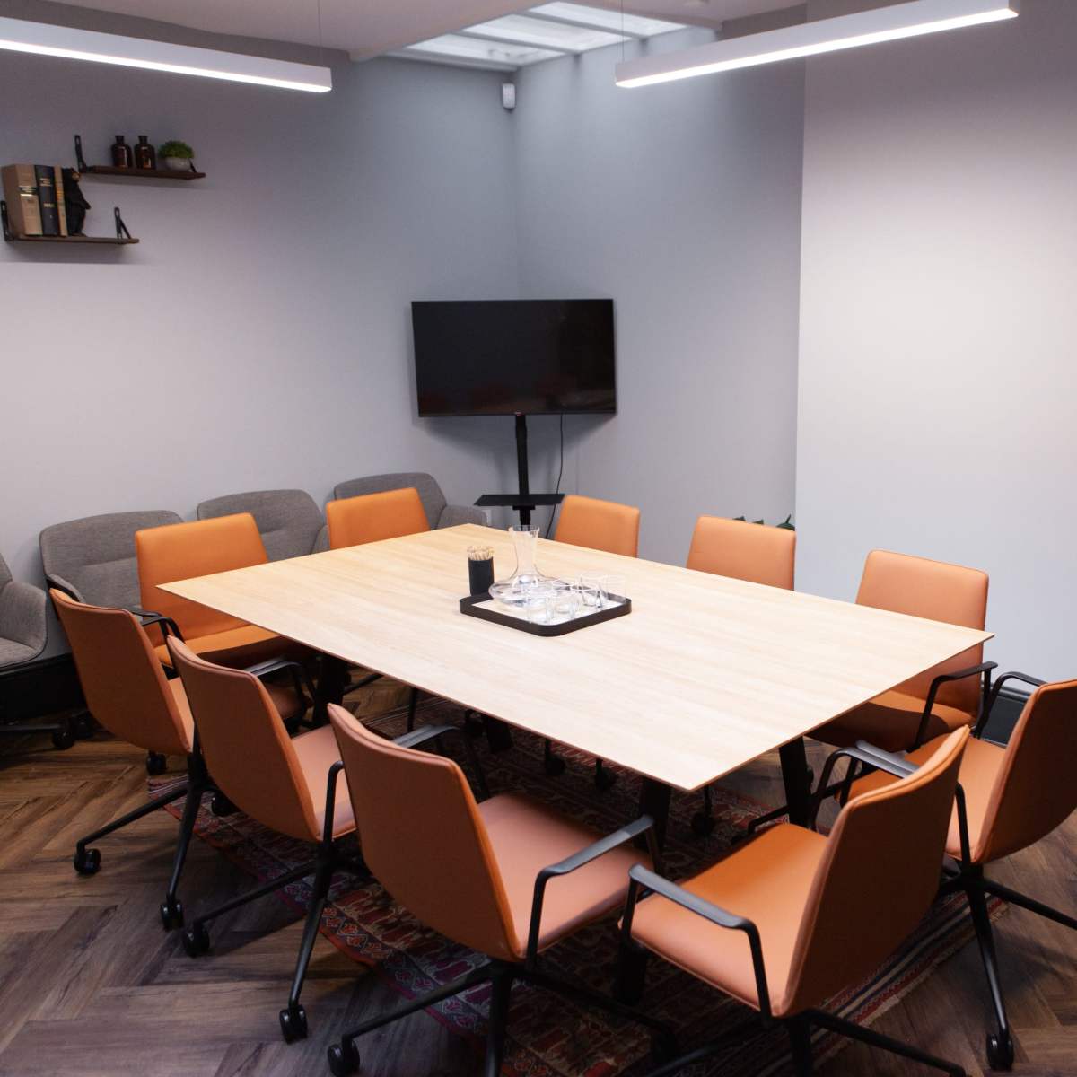 A modern conference room features a large wooden table surrounded by eight orange chairs, with a television and a floor lamp in the background.