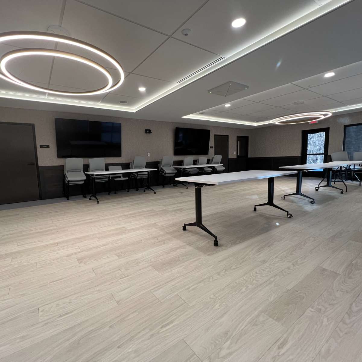 A modern meeting room with light wood flooring, two circular overhead lights, and tables arranged in the center.