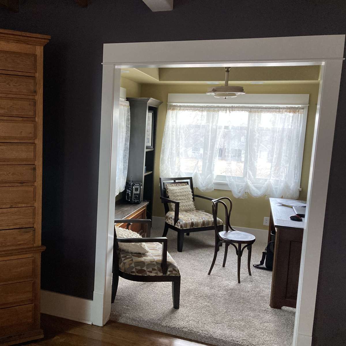 A tall, wooden chest of drawers stands beside an open doorway leading to a small, brightly lit sitting area.