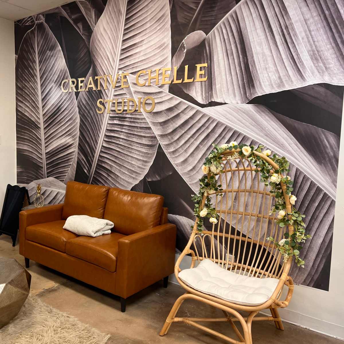 A cozy studio entrance with a brown leather sofa, a decorative chair, and a wall decorated with large leafy prints.