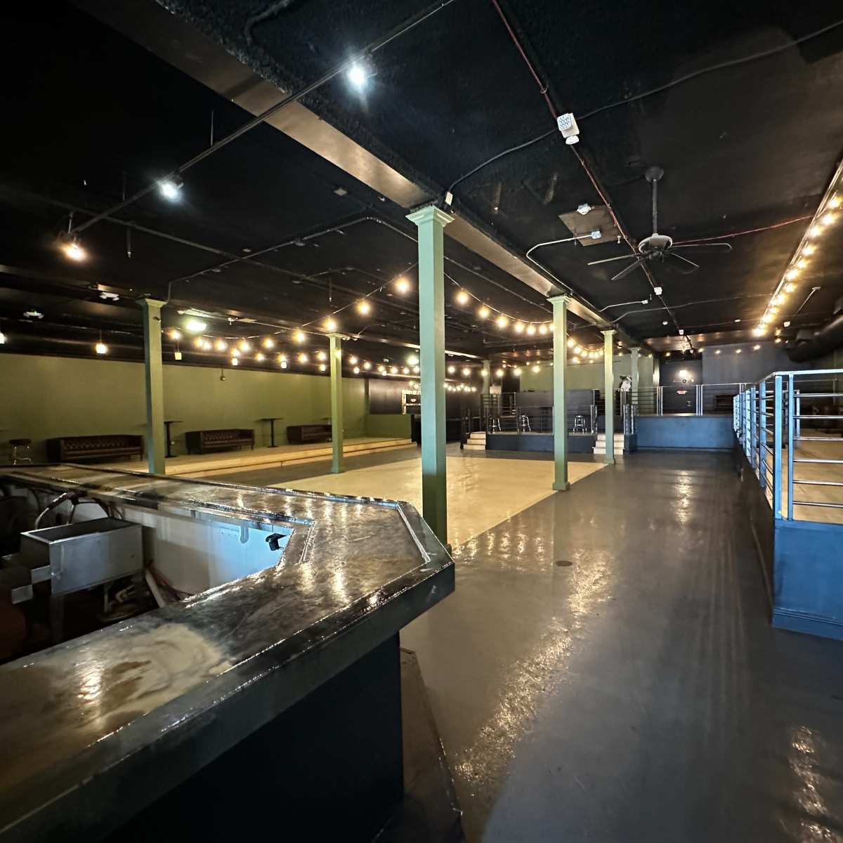 An empty interior of a venue with a polished floor, exposed lighting, and a bar area in the foreground.