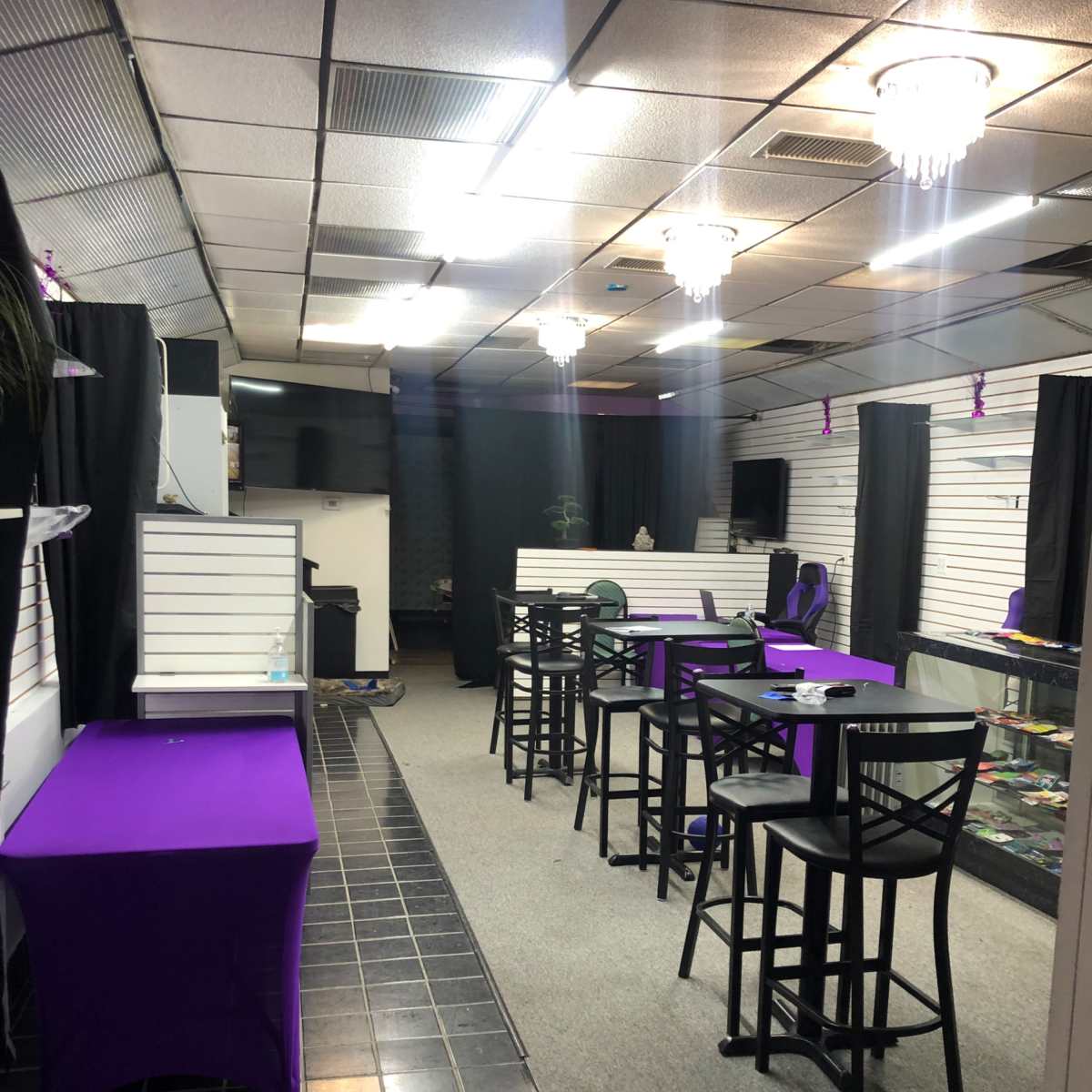 An empty retail space featuring black chairs and tables with purple tablecloths, displayed merchandise in glass cases, and illuminated by overhead lights.