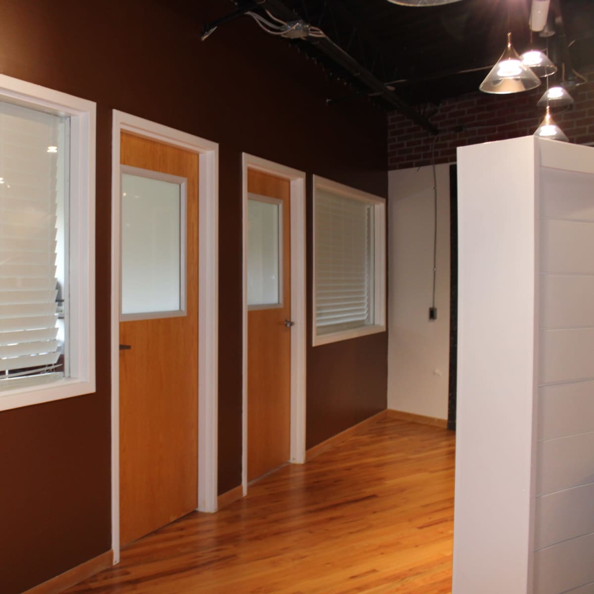 A corridor with two doors on the left, large windows with blinds, and wooden flooring.