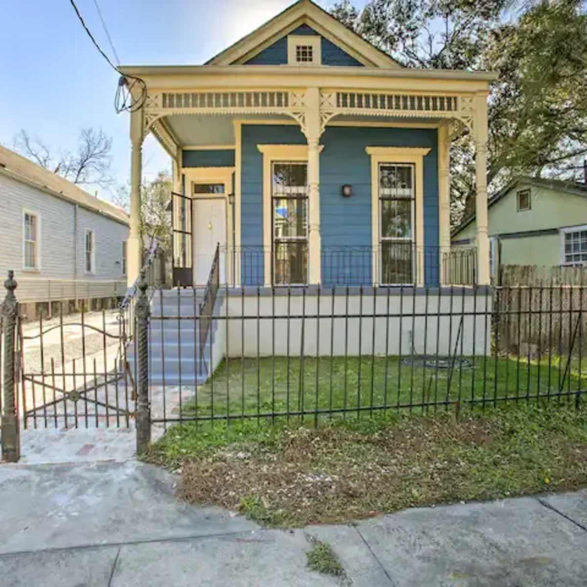 A blue wooden house with a front porch and decorative trim is surrounded by a black wrought-iron fence and is situated on a quiet street.