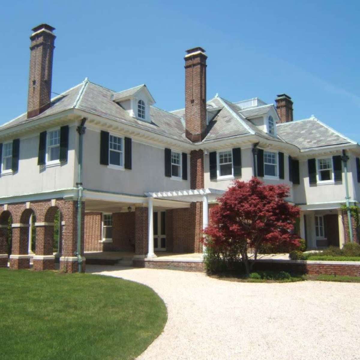 A large, two-story mansion with a circular driveway, visible brick chimneys, and a landscaped front yard.