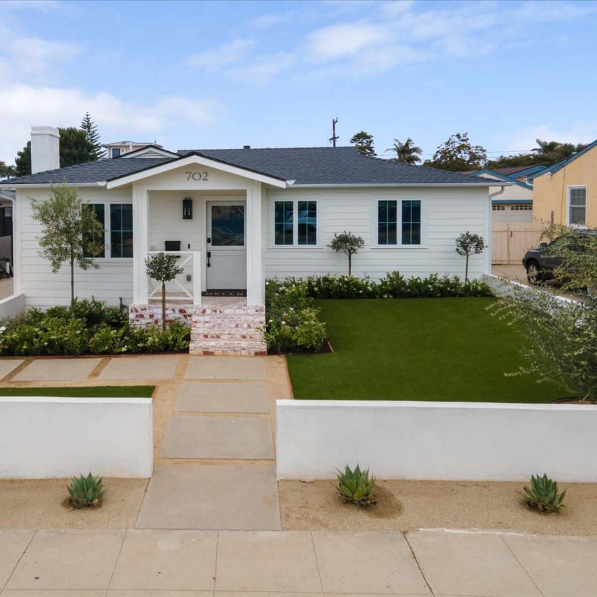 A single-story white house with a front lawn, bordered by a low wall and a walkway leading to the main entrance.