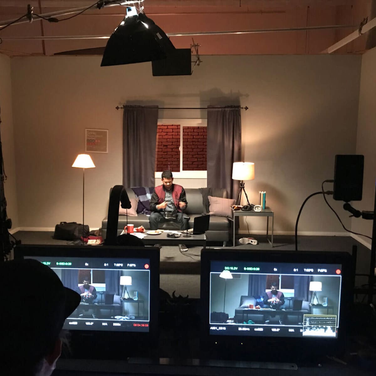 A man sits on a couch in a studio setting, with two monitors displaying the scene in front of him.
