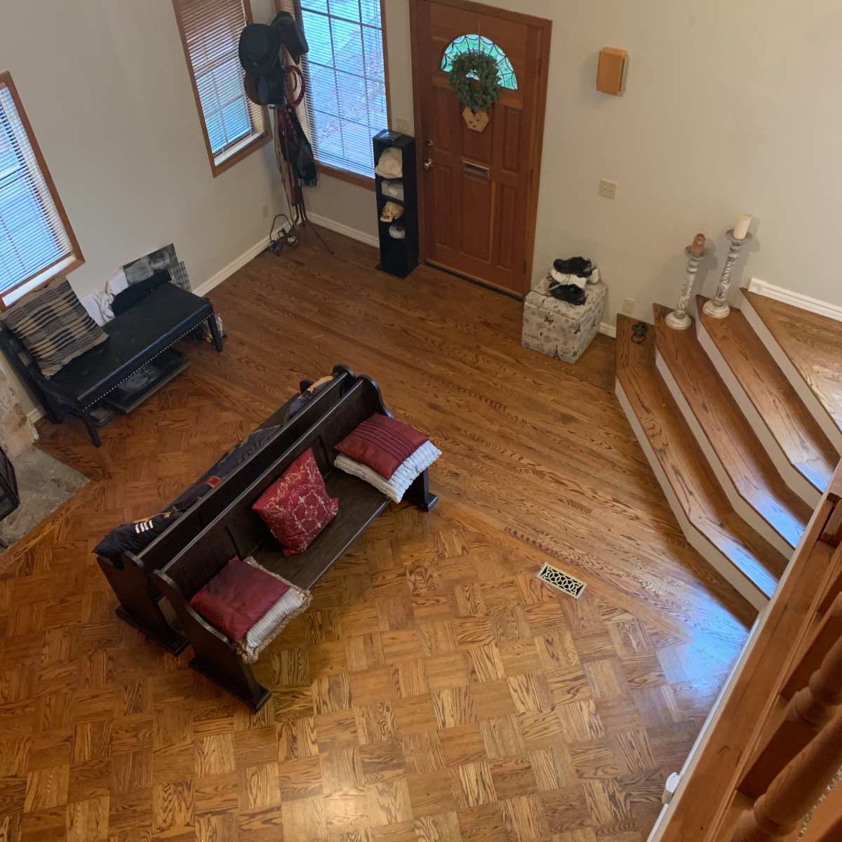 A living room with a wooden floor, a dark couch with red cushions, a staircase, and a front door with a wreath.