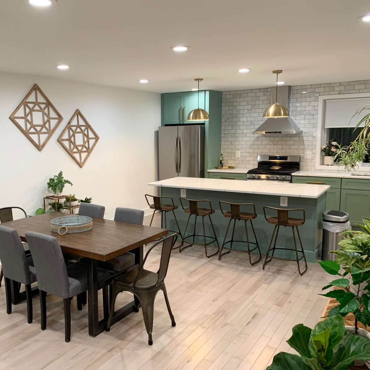 A modern kitchen and dining area features a wooden table surrounded by chairs, with a kitchen island, stainless steel appliances, and decorative wall art.