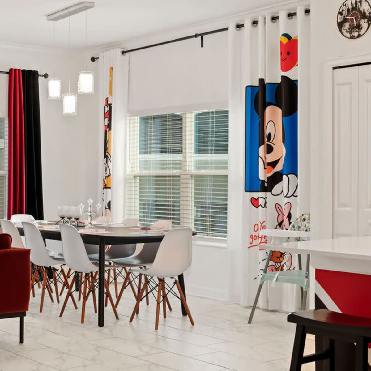 A dining area features a round table surrounded by white chairs, with a red sofa nearby and colorful decorations, including Mickey Mouse-themed curtains and cushions.