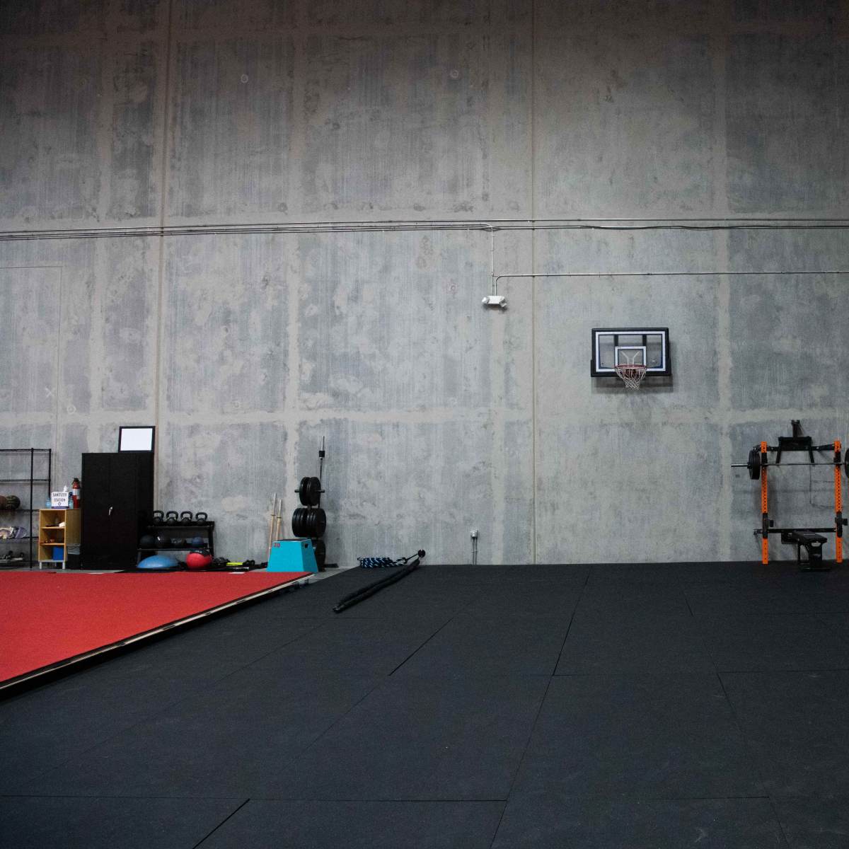 A spacious gym with a gray concrete wall, a basketball hoop, a punching bag on a stand, and exercise equipment along one side.