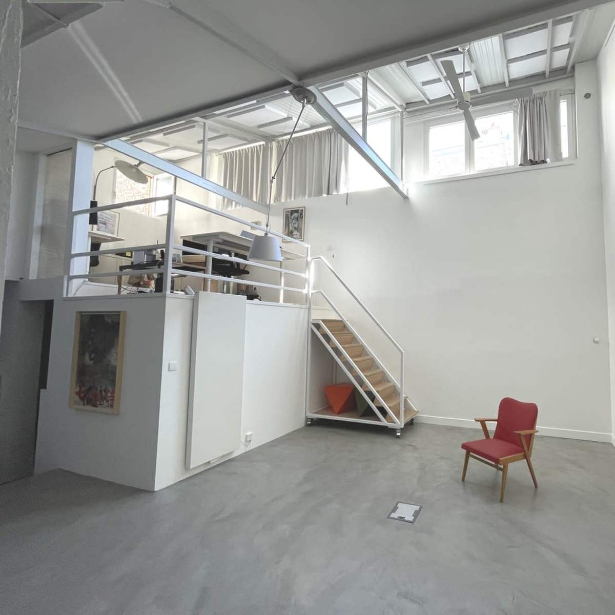 A spacious room with a wooden staircase leading to a loft area, a red chair on the floor, and large windows along the upper portion of the wall.