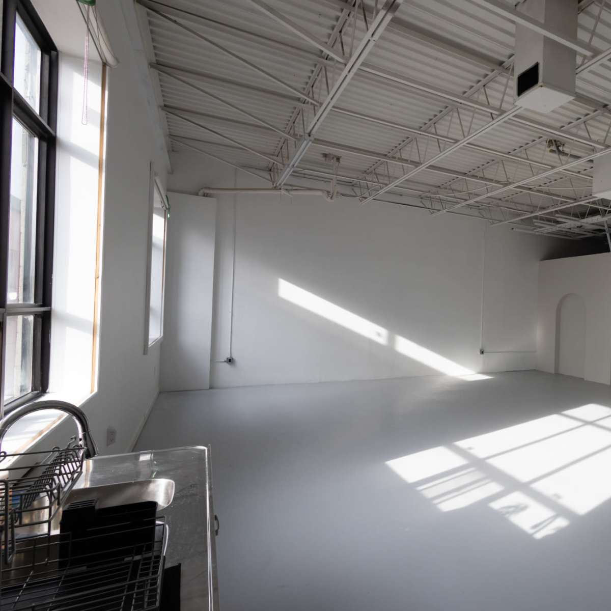 A spacious, well-lit studio with large windows and a white floor, featuring minimalistic archways on one wall.