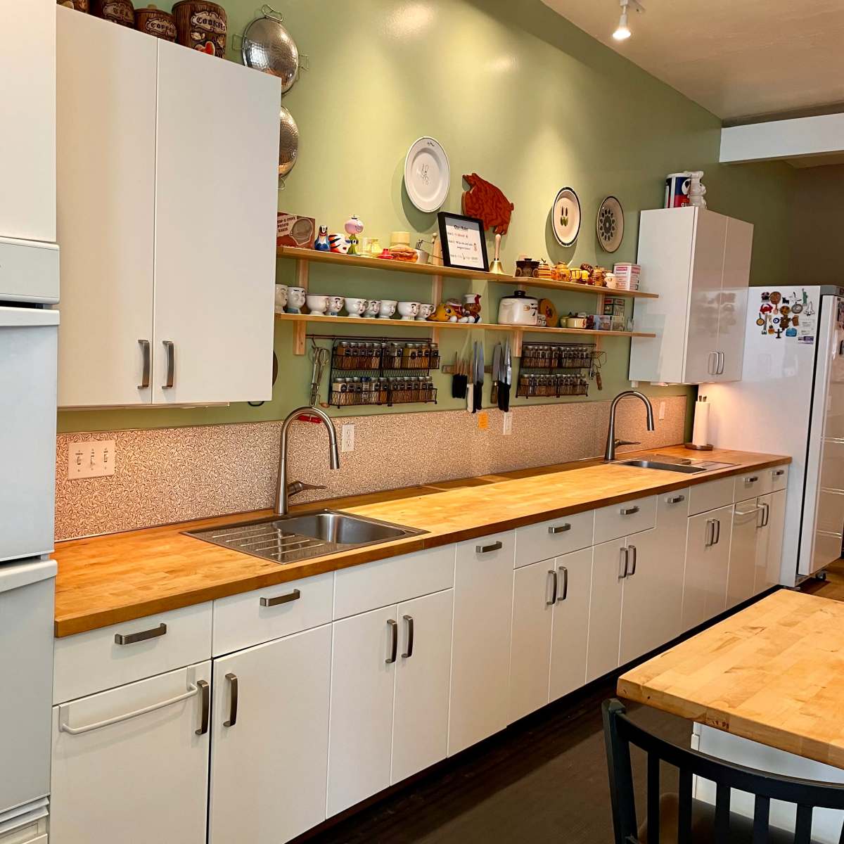 The kitchen features a long wooden countertop with double sinks, white cabinets, and organized shelves displaying dishes and spice jars against a green wall.