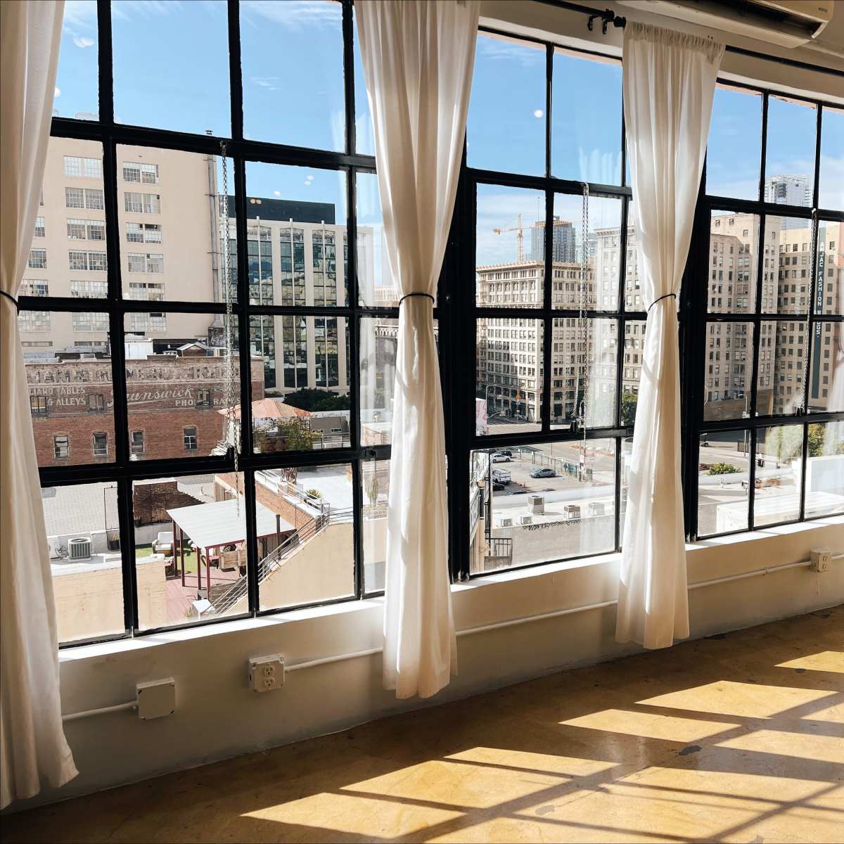 A sunlit interior with large windows and sheer curtains, revealing an urban view of buildings and streets outside.