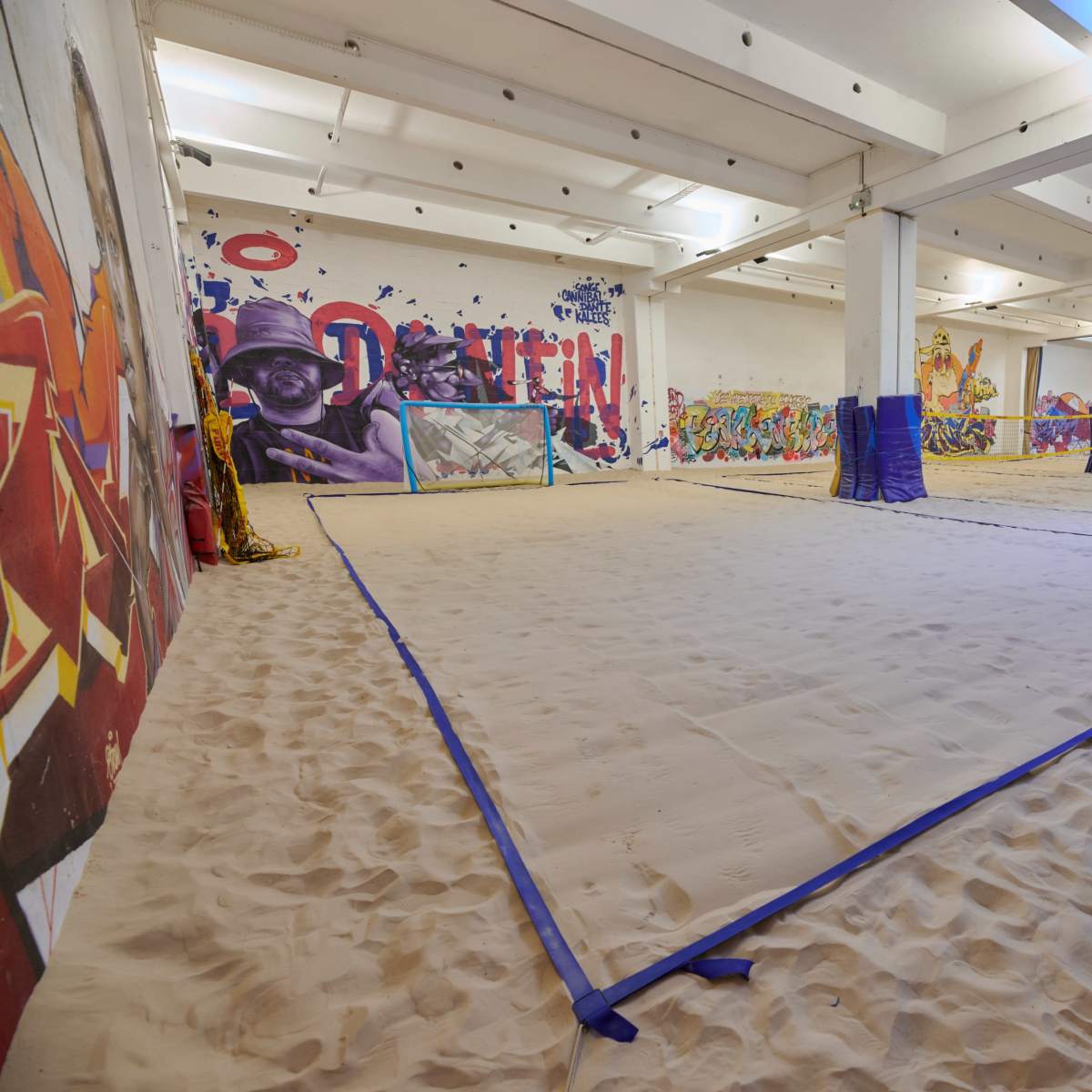 An indoor space with sand spread across the floor, marked by blue tarps and vibrant wall murals.