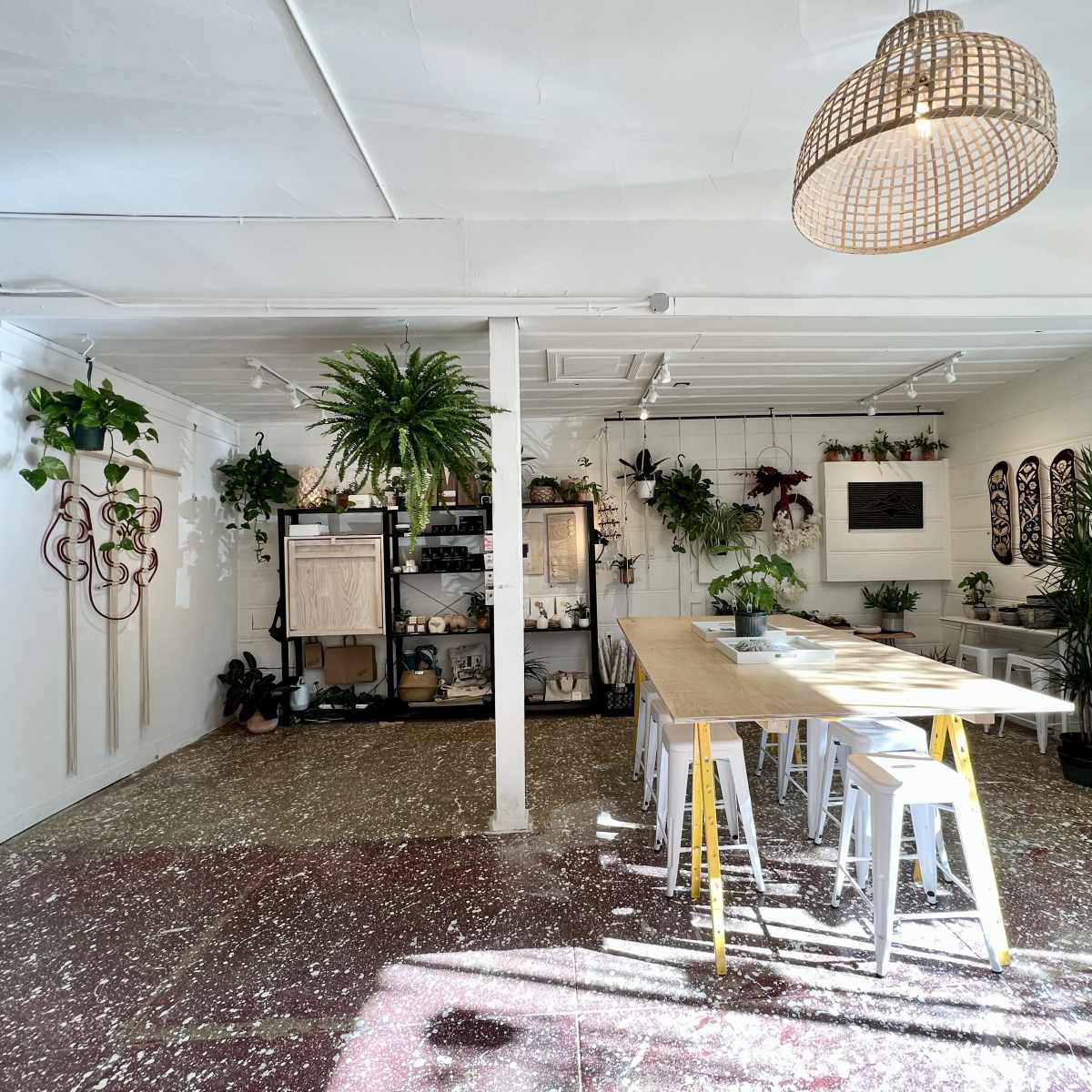 A bright interior space features a large wooden table surrounded by white stools, with various plants hanging and displayed around the room.