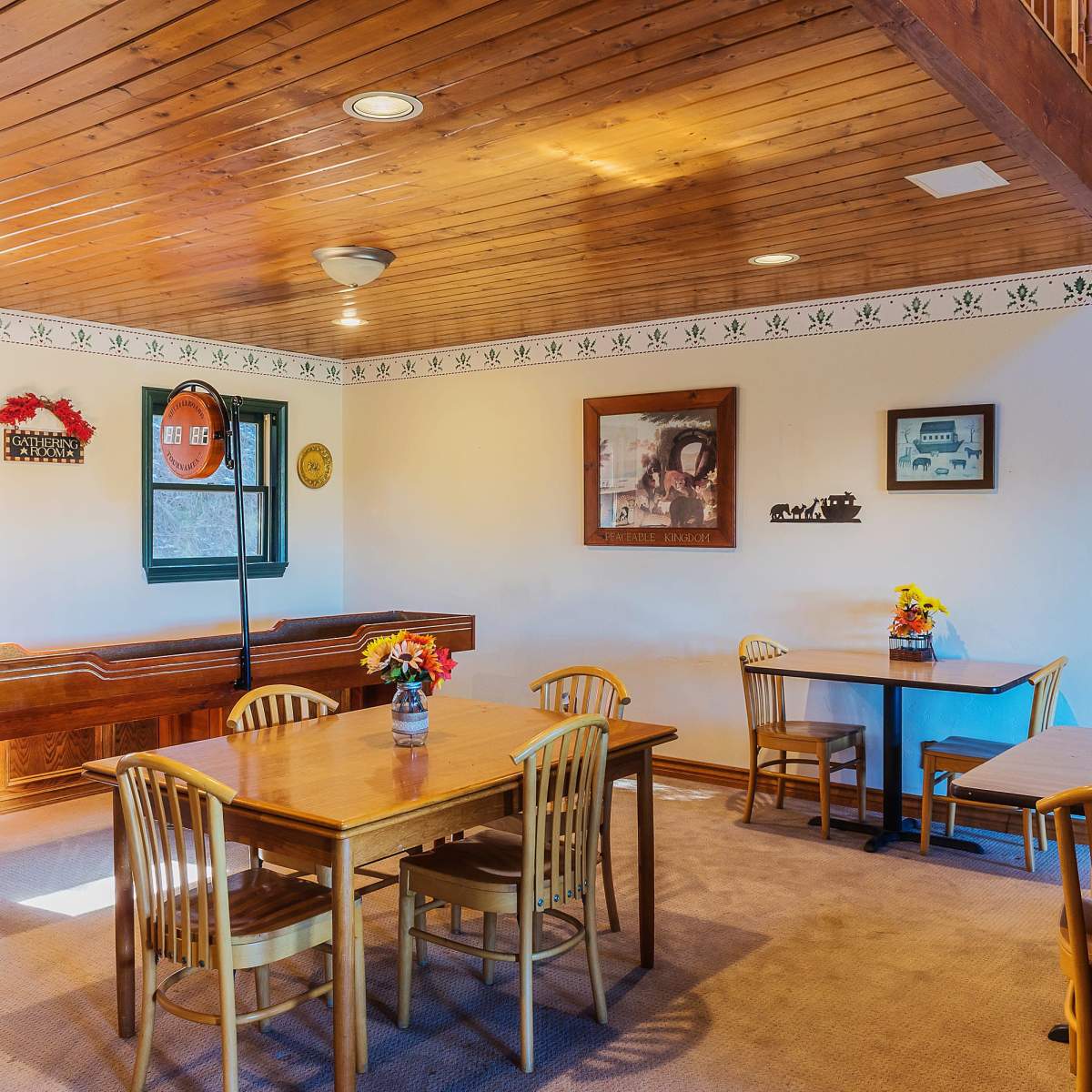 A cozy room features wooden beams, a stone wall, a pool table, and several wooden dining tables with chairs surrounded by decorative flower arrangements.