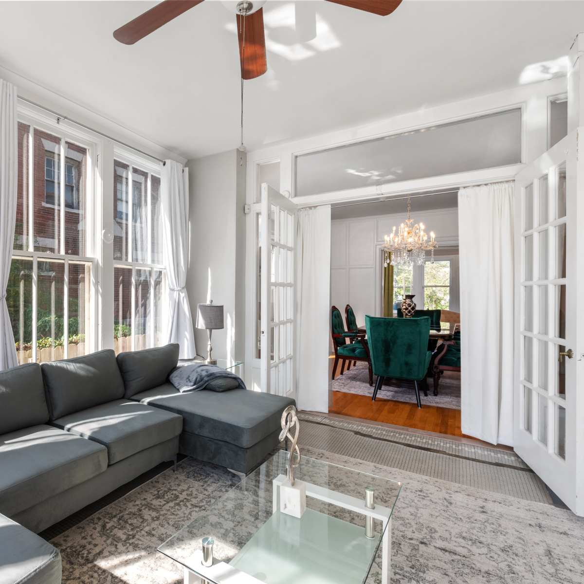 A modern living room with large windows, a gray sectional sofa, and open French doors leading to a dining area with green chairs.