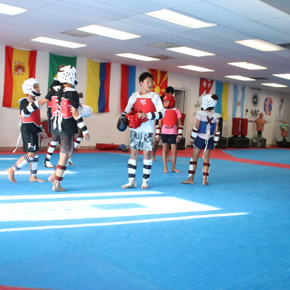 A group of children in protective gear practices martial arts in a brightly lit dojo decorated with various country flags.