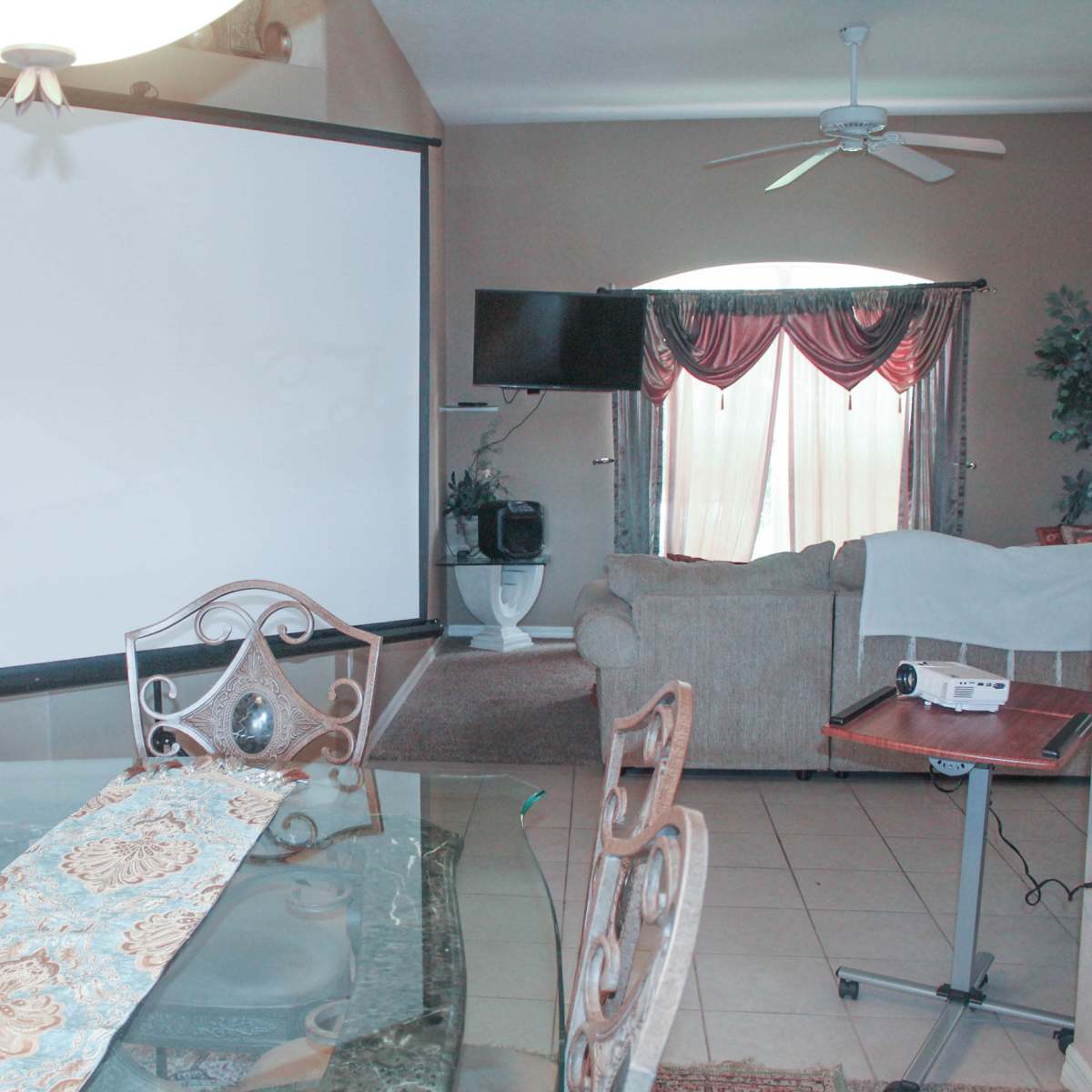 A dining area with a glass table and chairs adjacent to a living room featuring a projector screen and a TV.