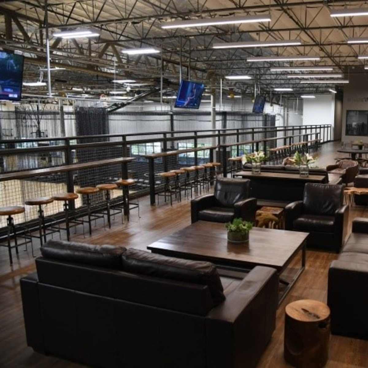 A spacious office area with leather couches, a wooden table, and a balcony overlooking a sports facility, complemented by large televisions mounted on the walls.