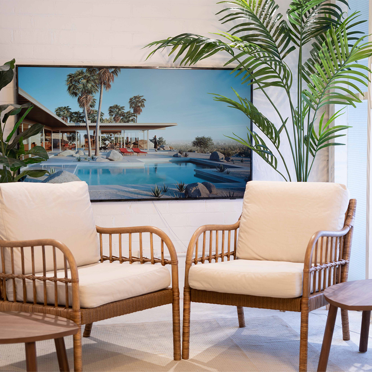 Two wicker armchairs with cream cushions, a small round wooden table, and a large framed landscape photograph of a modern poolside scene with palm trees.