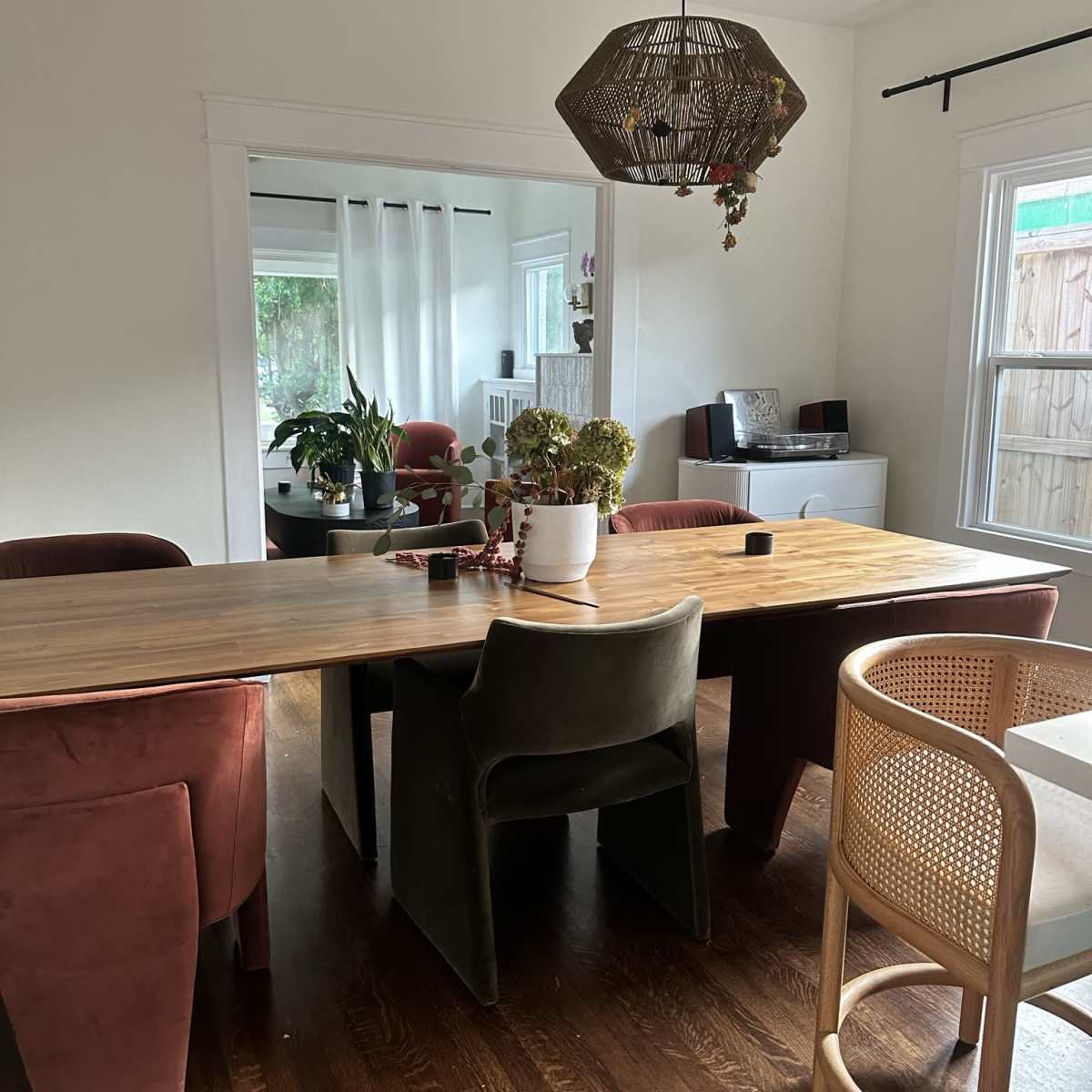 A dining area with a wooden table surrounded by four upholstered chairs and a rattan stool, with a hanging light fixture above and plants visible in the background.