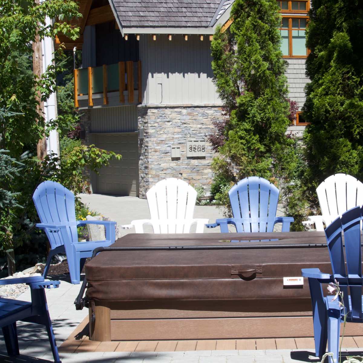 A row of blue and white Adirondack chairs surrounds a hot tub on a patio near a stone-faced house.