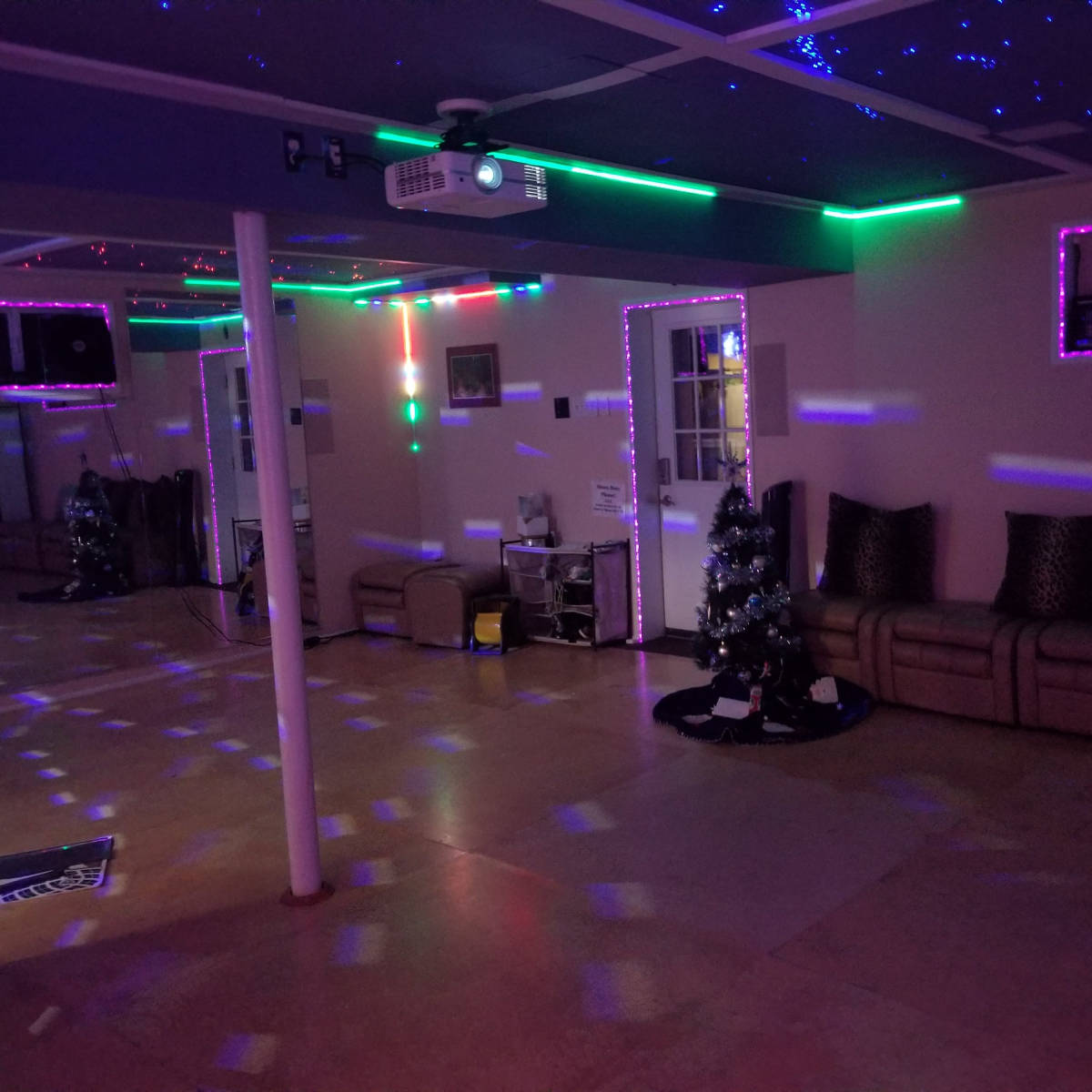 A spacious room with colorful lights, a projector on the ceiling, a Christmas tree in one corner, and a seating area with metallic pillows.