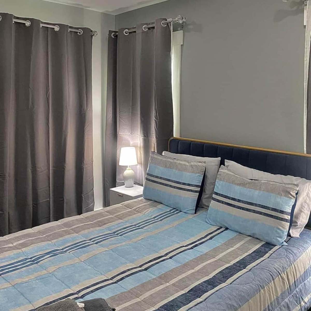 A bedroom with a bed featuring blue and gray striped bedding, two dark curtains covering the windows, and a small bedside lamp on a white nightstand.