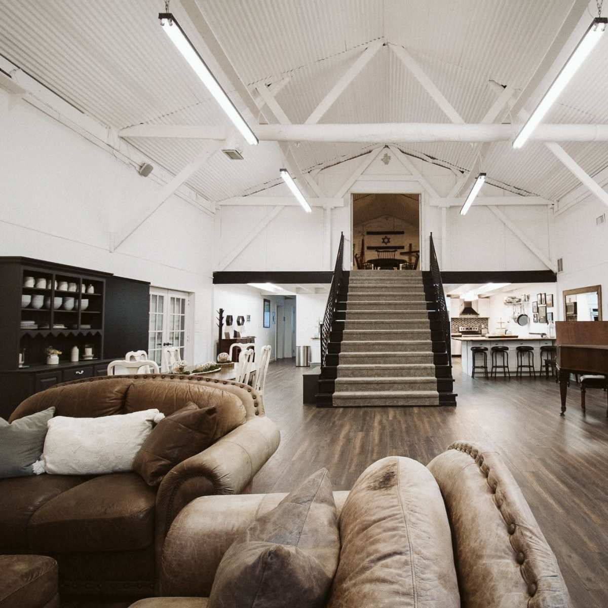 A spacious, open interior with high ceilings, featuring a staircase in the center that leads to an upper level, surrounded by various furniture arrangements.