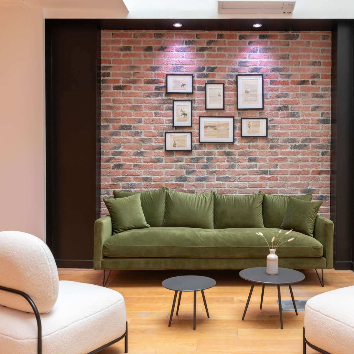 A green sofa is positioned against a brick wall adorned with framed artworks, with two round tables and two upholstered chairs in front.