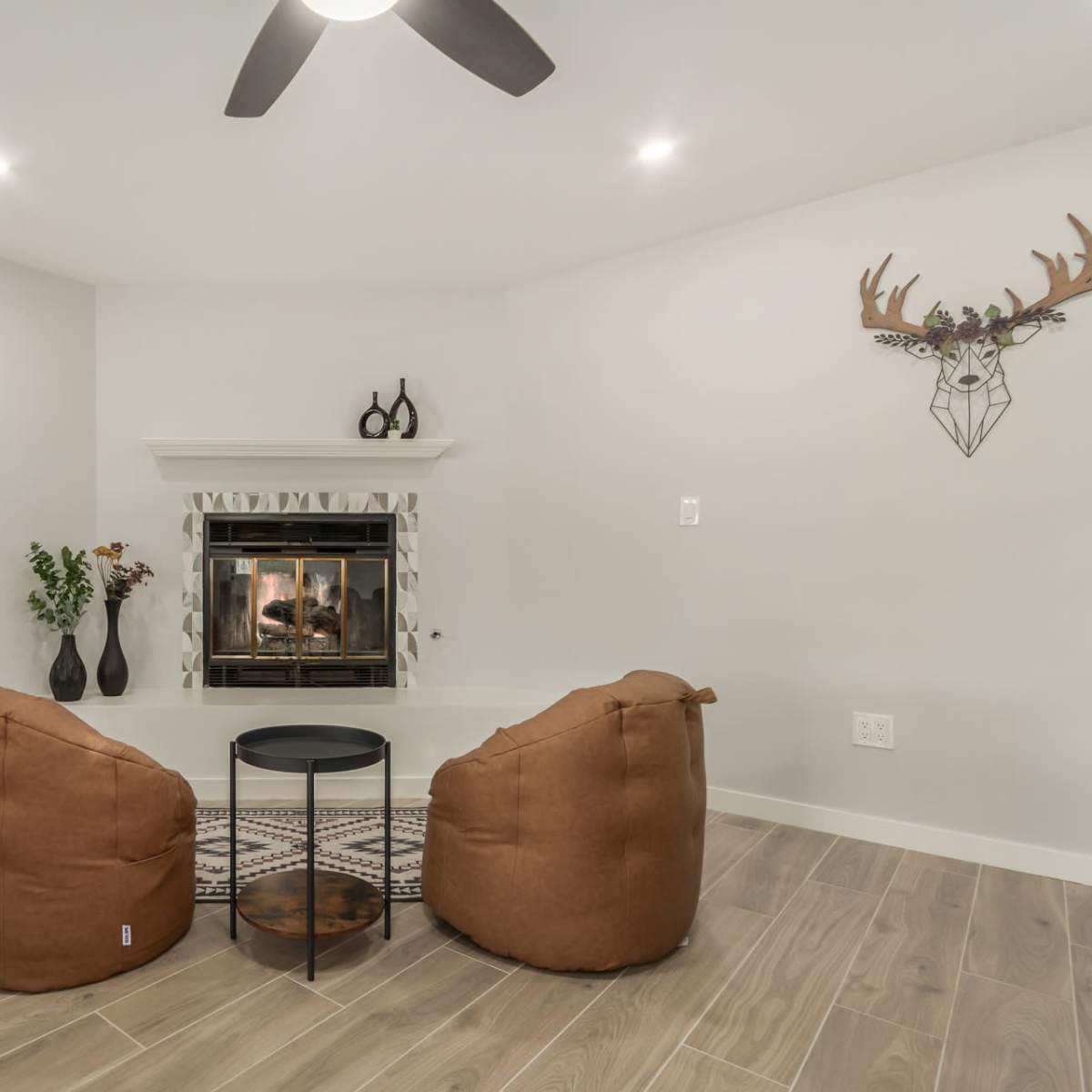 A cozy living room features two tan bean bag chairs facing a fireplace, with a small black table in between and decorative elements on the walls.
