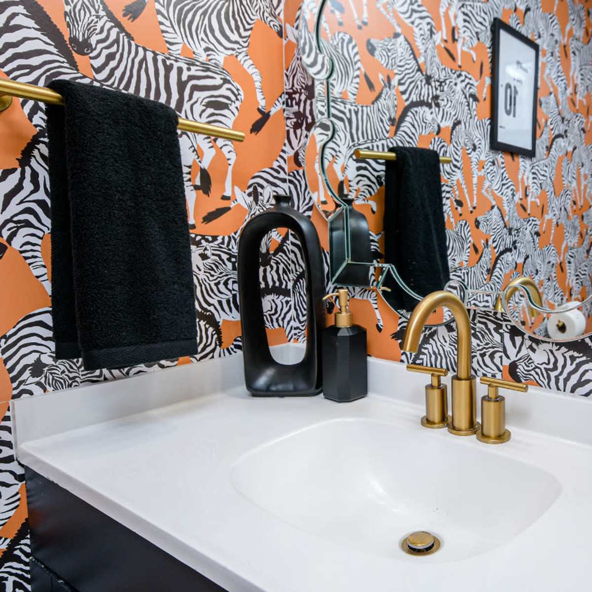 The bathroom features a white sink with a gold faucet and black accessories against a vibrant zebra-striped wallpaper.