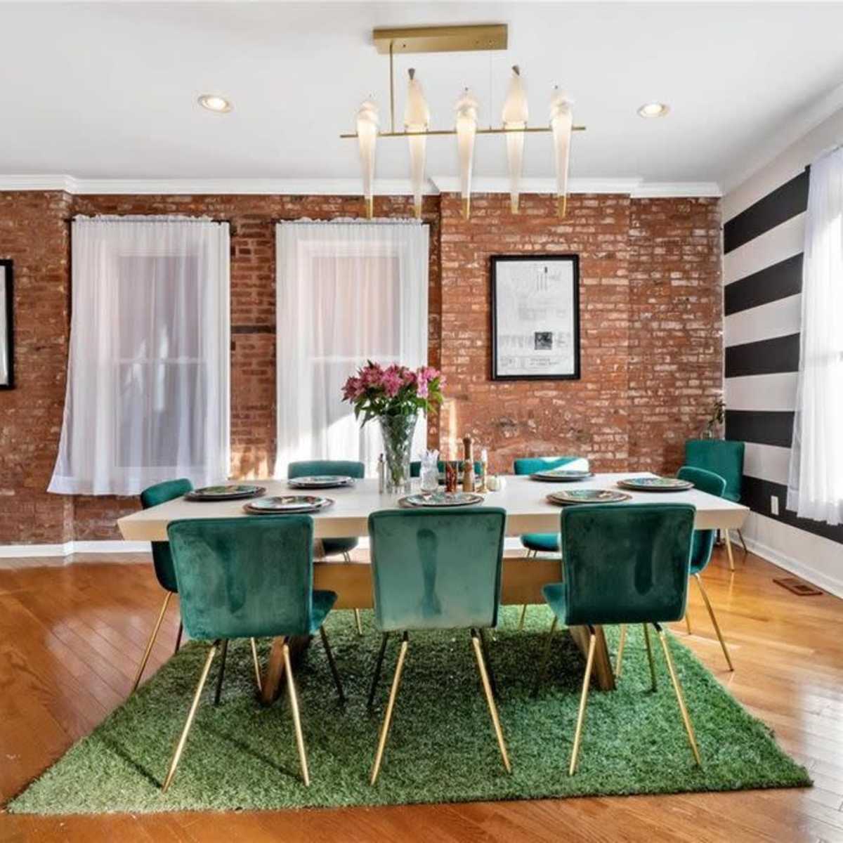 A dining room features a large table surrounded by green velvet chairs on a grass-like rug, with exposed brick walls and striped accents.