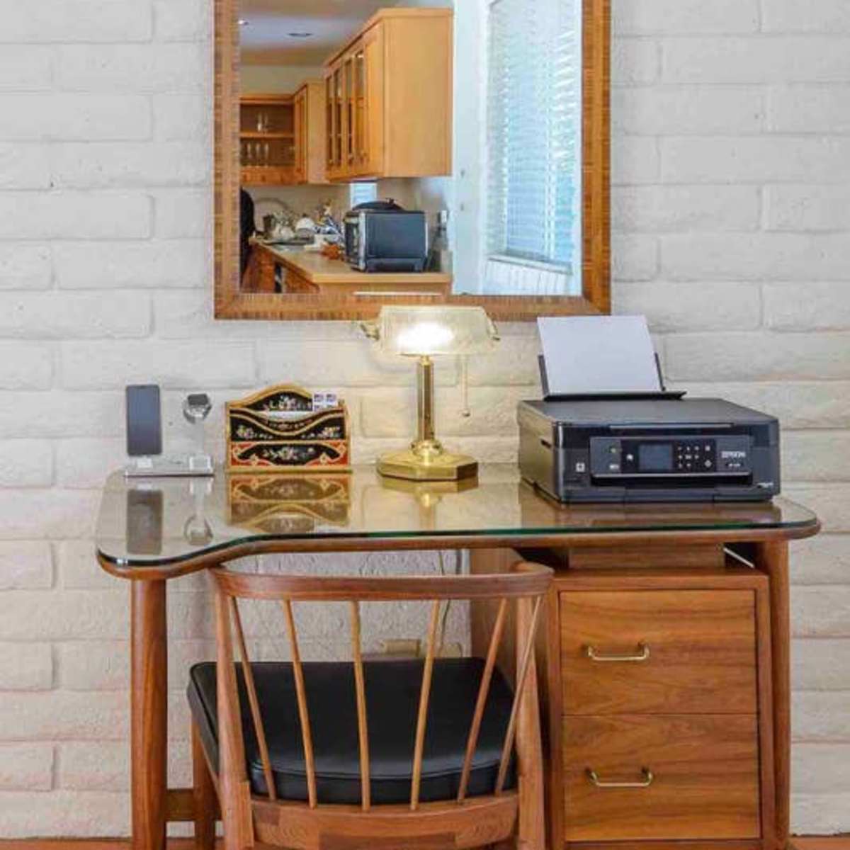 A wooden desk with a printer, lamp, and drawer is placed against a white brick wall, reflecting a kitchen area in a nearby mirror.