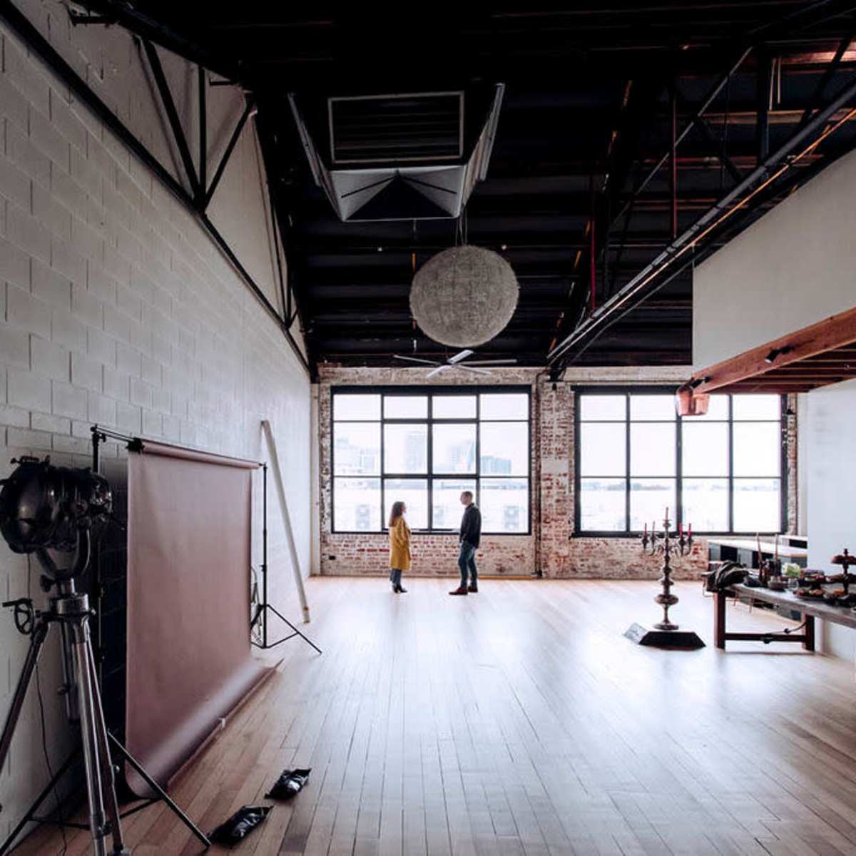 Two people stand in a spacious, industrial-style room with large windows and wooden flooring, surrounded by photography equipment and furnishings.
